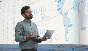 Stock image of Indian Male Logistics Expert Holding Laptop Computer And Analyzing World Map On Big Digital Screen In Monitoring Office. 